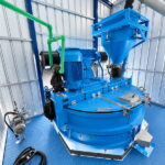 Máquina azul con tubos verdes, un motor lateral y una tolva de descarga, instalada dentro de una caseta de chapa metálica