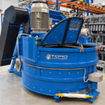 Máquina circular azul con motor superior, chapas negras plegables y el rótulo "TORO Machinery" en el cuerpo, junto a moldes metálicos apilados