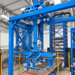 Mecanismo azul con motor y cadena, sujetando con una pinza metálica un conjunto de bloques grises sobre una plataforma de madera
